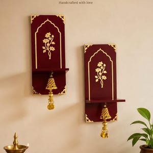 May include: Two rectangular maroon wall shelves featuring gold-coloured floral motifs and arched borders. Each shelf includes a hanging gold-coloured bell ornament with dangling circular accents. The text Mahi craft studio Handcrafted with love is visible.
