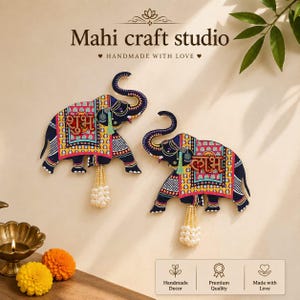 May include: Two decorative wall hangings shaped like elephants, featuring dark blue bodies with colourful geometric patterns in red, yellow, and white. Each elephant displays Hindi text on its side and includes a dangling tassel of white beads.