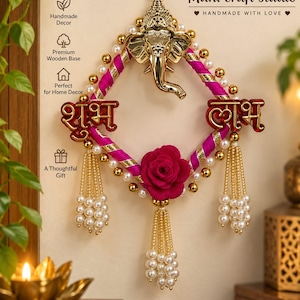 May include: A diamond-shaped wall hanging featuring a gold-toned Ganesha figure at the top, magenta fabric-wrapped borders, and white pearl accents. The Hindi text Shubh Labh is displayed in red, with a large magenta fabric rose and pearl tassels.