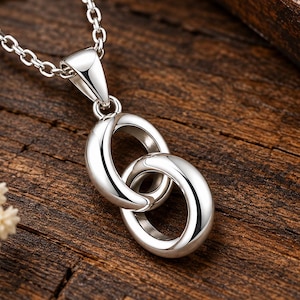 Puede incluir: Un collar con colgante de tono plateado pulido que presenta dos anillos ovalados entrelazados suspendidos de una cadena delicada. La superficie metálica es lisa y reflectante, mostrando un diseño limpio con una argolla que conecta el colgante a la cadena.