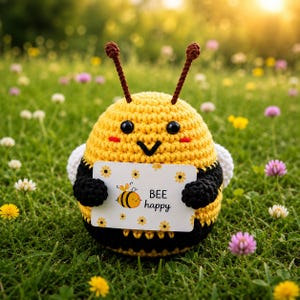 May include: A round, crocheted bee plush toy with yellow and black horizontal stripes, two brown antennae, and black safety eyes. It holds a white rectangular card featuring a small bee illustration and the printed text "BEE happy".