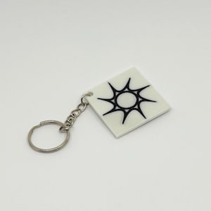 May include: A square white keyring featuring a black graphic of a stylised sun with eight sharp, pointed rays. The pendant is attached to a silver-toned metal split ring by a short chain, providing a simple and functional design for keys.