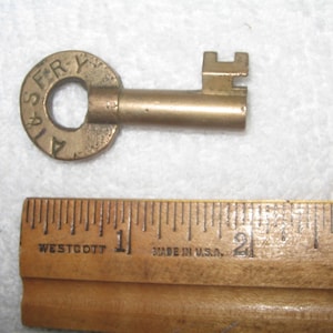 May include: A brass-colored metal key with a circular bow featuring the stamped letters A & S FRY. The key is positioned above a wooden ruler, showing a length of approximately 2.5 inches from the bow to the bit of the key.