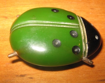 May include: A beetle-shaped brooch featuring a smooth, rounded green body with five black circular spots and a distinct black head section. The piece includes small metallic protrusions on the sides and a central horizontal line.