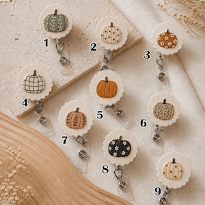 May include: Nine circular badge reels with scalloped edges, each featuring a unique pumpkin design. Designs include a green pumpkin with vines, a white pumpkin with hearts, an orange pumpkin with daisies, a plaid pumpkin, a solid orange pumpkin, a polka dot pumpkin, a tan pumpkin with vines, a black pumpkin with daisies, and a white pumpkin with flowers.