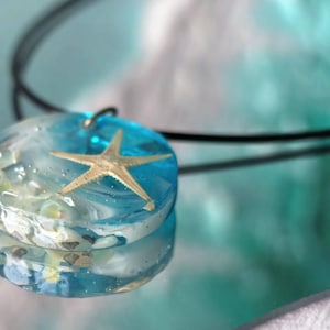 Könnte beinhalten: Eine runde Anhängerkette mit einer transparenten Harzscheibe in hellblauem und weißem Wirbelmuster, die einen kleinen beigen Seestern und winzige weiße Muschelfragmente enthält, befestigt an einer dünnen schwarzen Kordelkette.