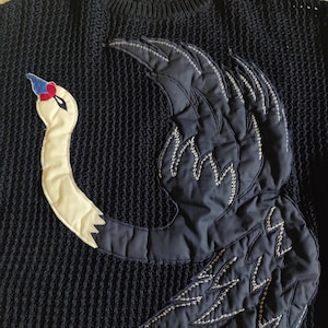 May include: A dark navy blue knit sweater featuring a large, quilted appliqué of a swan. The swan has a cream-colored neck, a blue and red beak, and dark navy wings with white decorative stitching. An Escada brand label is at the neckline.