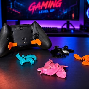 May include: A black video game controller with orange paddle attachments installed on the back. Several additional sets of paddle attachments in bright orange, teal, pink, and black are arranged in front of the controller on a dark surface.