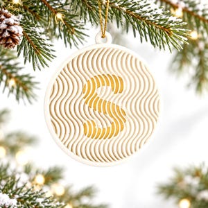 May include: A circular white ornament featuring a layered, wavy line pattern with a prominent gold letter S at the center. The ornament is suspended by a thin gold string and displays a dimensional design with cutouts.