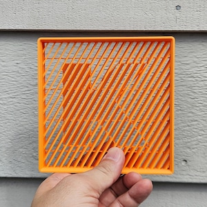 May include: A square orange coaster featuring a diagonal slat pattern and a central cutout in the shape of the letter K. The object has a raised border frame and a grid-like structure, constructed from a solid, matte-finish material.