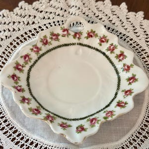 May include: A white porcelain dish with a scalloped edge and a single handle. The rim features a repeating pattern of small pink roses and green leaves, enclosed by a dark green laurel leaf border. The center of the dish is plain white.