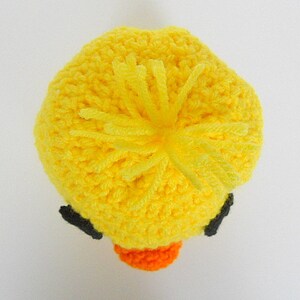 May include: A yellow crocheted duck hat with black eyes and an orange beak. The hat has a fluffy yellow pom-pom on top.