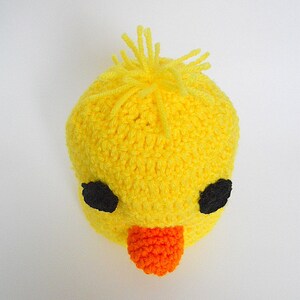 May include: A yellow crocheted baby hat with black eyes and an orange beak. The hat has a fluffy yellow top.