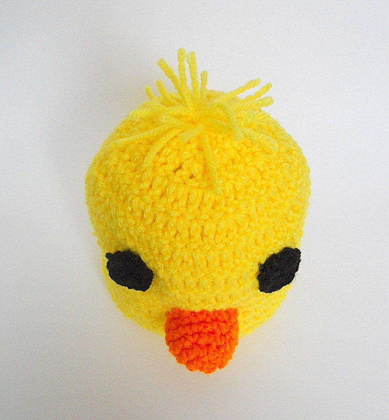 May include: A yellow crocheted baby hat with black eyes and an orange beak. The hat has a fluffy yellow top.