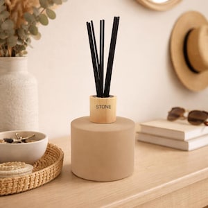 May include: A cylindrical beige reed diffuser with a light wood collar featuring the word STONE printed in black. The diffuser contains several thin black reeds extending upwards from the centre of the vessel, which has a smooth, matte finish.