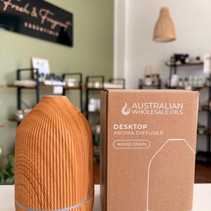 May include: A wood grain aroma diffuser with a vertical ribbed texture and a power button at the base, positioned next to its cardboard packaging. The box features the text Australian Wholesale Oils, Desktop Aroma Diffuser, Wood Grain, USB Powered, Multi-colour LED Light, and 100ml Capacity.
