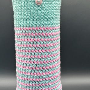 May include: A light blue and pink striped crochet water bottle sleeve featuring a small pink button at the top center. The sleeve is fitted around a white Owala water bottle with a yellow and orange lid, showcasing a textured yarn stitch pattern.