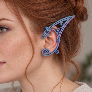 May include: An iridescent blue and purple ear cuff featuring an intricate filigree design with swirling patterns that extend upward into a pointed shape, wrapping around the outer edge of the ear with small metal loops for secure placement.