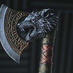 Könnte beinhalten: Eine dekorative Axt mit einem skulptierten Wolfskopf an der Seite der Klinge. Die Klinge hat ein metallisches Finish mit komplizierten eingravierten Mustern. Der Griff ist mit braunem Material mit Kreuzschraffur und metallischen Akzenten umwickelt.