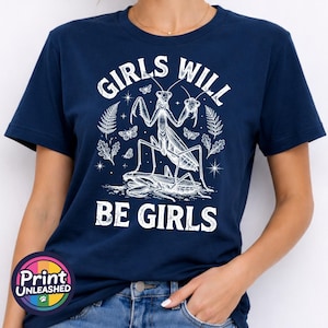 May include: A navy blue short-sleeved t-shirt featuring a white graphic print of a praying mantis standing over another insect. The text "GIRLS WILL BE GIRLS" is printed in a distressed white font above and below the central insect illustration.