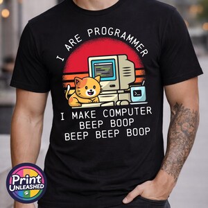 May include: A black t-shirt featuring a graphic of an orange cat sitting next to a vintage computer. The text reads I ARE PROGRAMMER I MAKE COMPUTER BEEP BOOP BEEP BEEP BOOP. The design includes a red sun background with horizontal stripes.