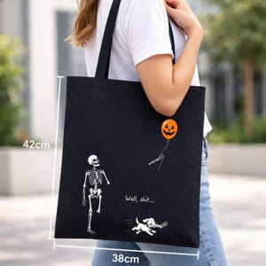 May include: A black tote bag measuring 42cm by 38cm featuring a white skeleton graphic, a dog, and an orange jack-o'-lantern balloon. The skeleton holds the balloon string, and the text "Well, shit..." is printed above the dog.