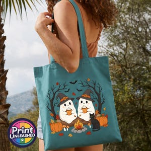 May include: A teal tote bag featuring a graphic of two white ghosts wearing witch hats, sitting by a campfire with a black cat and pumpkins. The ghosts hold orange mugs. The scene includes bare trees, falling autumn leaves, and flying bats.