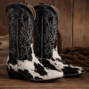 Puede incluir: Un par de botas de estilo western con estampado de piel de vaca en blanco y negro en el pie y una caña de cuero negro. La parte superior de la caña está decorada con intrincadas costuras blancas en forma de llama y un tacón.
