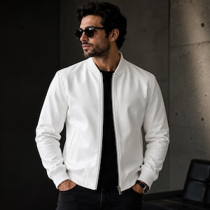 May include: A white bomber jacket featuring a ribbed collar, cuffs, and hem, with a full-length front zipper closure. The jacket is worn over a plain black t-shirt and paired with dark trousers, complemented by black sunglasses and a wrist watch.