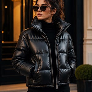 May include: A black puffer jacket with a high collar, horizontal quilted stitching, and a full-length silver zip closure. The jacket features a smooth, semi-gloss finish and long sleeves, worn over a black turtleneck jumper and black trousers.