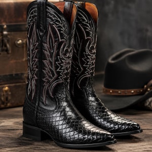 May include: A pair of black cowboy boots featuring a textured snakeskin-patterned leather foot and a smooth leather shaft with intricate white and red decorative stitching, a pointed toe, and a stacked heel with a pull tab on each side.