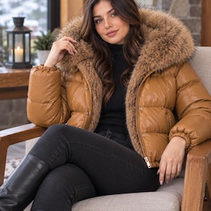 Könnte beinhalten: Eine beige Steppjacke mit einer großen Kapuze mit dickem braunem Kunstfellbesatz, langen Ärmeln mit elastischen Bündchen, einem Frontreißverschluss und einem horizontalen Steppmuster. Die Jacke wird über einem schwarzen Oberteil mit Stehkragen getragen.
