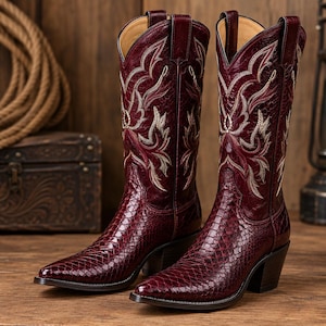 May include: A pair of deep burgundy cowboy boots featuring a textured snakeskin-patterned leather foot and a tall shaft adorned with intricate cream and gold-toned embroidery. The boots have a pointed toe, a stacked heel, and pull tabs at the top.