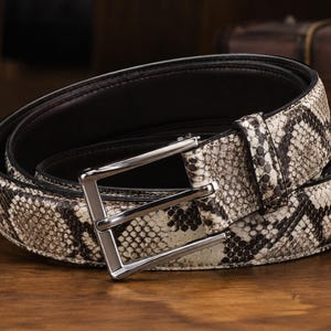 May include: A belt featuring a textured snakeskin pattern in shades of cream, beige, and dark brown. The belt includes a polished silver-tone metal rectangular buckle and a matching leather keeper loop, with a smooth black interior lining.