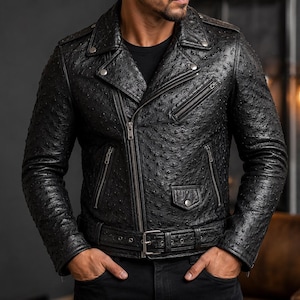 Può includere: Una giacca da moto in pelle nera con motivo testurizzato in pelle di struzzo, dotata di cerniera frontale diagonale, finiture in metallo argentato, tasche multiple con zip, bavero con bottoni a pressione e una cintura regolabile in vita.