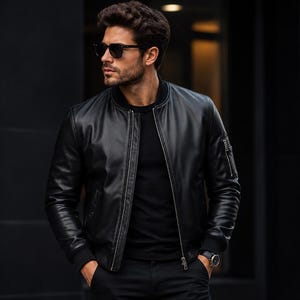 May include: A black leather bomber jacket featuring a front zip fastening, ribbed cuffs, and a small zipped utility pocket on the left sleeve. The jacket is worn over a plain black t-shirt, paired with black trousers and a black wristwatch.