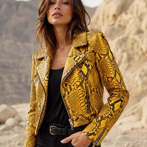 Puede incluir: Una chaqueta motera con estampado de piel de serpiente en amarillo y negro, con solapas, cremalleras metálicas plateadas en la parte delantera y en los puños, y un corte corto. La chaqueta se lleva sobre una camiseta negra lisa y pantalones negros con cinturón.