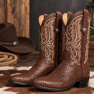 Op de afbeelding: Een paar bruine cowboylaarzen met een gestructureerd slangenleerachtig patroon op het leer en ingewikkeld crèmekleurig borduurwerk op de schacht. Elke laars heeft een treklipje met een klein zilverkleurig metalen detail aan de bovenkant.