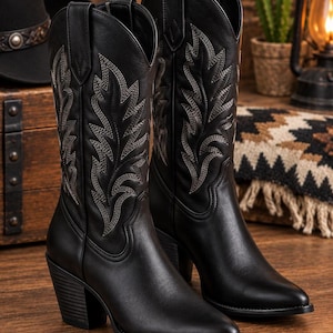 May include: A pair of black leather cowboy boots featuring a pointed toe, a stacked heel, and intricate light-colored stitching in a leaf-like pattern along the tall shaft. The boots have pull tabs at the top and a smooth, matte finish.