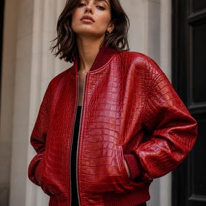 May include: A vibrant red bomber jacket featuring a textured crocodile-embossed pattern. The jacket includes a silver-toned front zip closure, ribbed knit cuffs, a ribbed collar, and a ribbed hemline, providing a structured and fitted appearance.