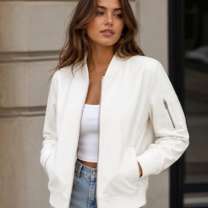 May include: A white bomber jacket featuring a front zipper closure, ribbed collar, cuffs, and hem, along with a zippered utility pocket on the left sleeve. The jacket is worn over a white tank top and light blue denim jeans with a visible metal button.