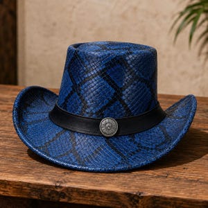 Puede incluir: Un sombrero de vaquero azul con un patrón texturizado de piel de serpiente en tonos azules y negros. El sombrero incluye una banda negra lisa que rodea la copa, decorada con un adorno circular de metal plateado en el centro de la parte frontal.