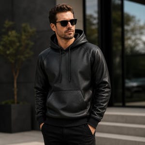 Peut inclure: Un sweat à capuche en cuir noir doté d'une poche kangourou sur le devant, d'une capuche avec cordons de serrage noirs assortis, et de poignets côtelés aux manches et à l'ourlet. Le vêtement présente une texture lisse et une coupe structurée.