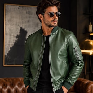 May include: A forest green leather bomber jacket featuring a ribbed collar, cuffs, and hem. The jacket has a central silver zipper closure and a small zippered utility pocket on the upper left sleeve. It is worn over a plain black shirt.