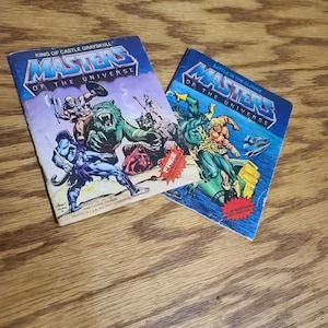 May include: Two vintage Masters of the Universe comic books titled King of Castle Grayskull and Battle in the Clouds. The covers feature colorful illustrations of He-Man and other characters in action, with red starburst text stating Free Illustrated Book With Purchase.