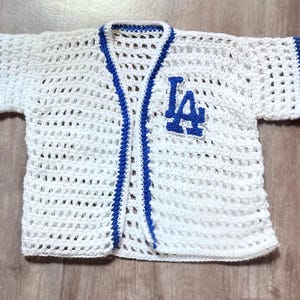 May include: A white crochet cardigan with short sleeves, featuring blue trim along the edges and sleeves. The front left side displays the blue embroidered letters LA. The garment is constructed with an open, airy stitch pattern throughout the body.