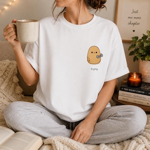 Puede incluir: Una camiseta blanca de manga corta con un pequeño gráfico en el pecho izquierdo de una patata de color marrón claro levantando una mancuerna gris. Debajo de la patata, la palabra "trying." está impresa en una fuente negra sencilla.