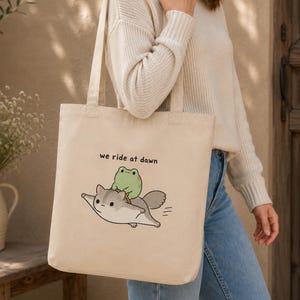 May include: A beige canvas tote bag featuring a graphic illustration of a small green frog riding on the back of a flying squirrel. Above the characters, the text "we ride at dawn" is printed in a simple black sans-serif font on the front panel.