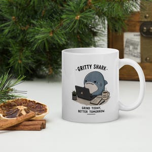 May include: A white ceramic mug featuring a cartoon illustration of a shark working on a laptop. The text reads GRITTY SHARK and GRIND TODAY, BETTER TOMORROW. The shark sits at a desk with a small plant, a to-do list, and a coffee mug.