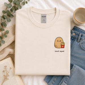 May include: A cream-coloured crew neck t-shirt featuring a small graphic of a brown potato character holding a red container of french fries with a yellow heart. The text "snack squad" is printed in black lowercase letters below the graphic.